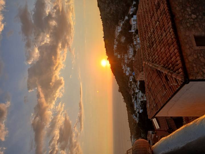 Golden sunset over a coastal hillside village with tiled rooftops and the sea on the horizon.