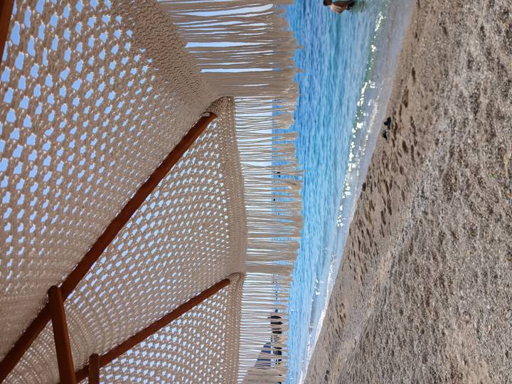 View of a sandy beach and calm sea framed by the fringe of a woven parasol.