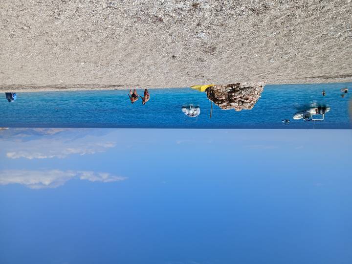Open pebble beach with swimmers, anchored boats and a clear blue sea against a mountain backdrop.