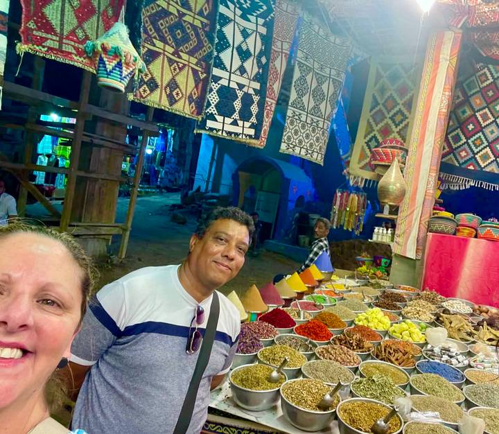 Evening market scene with colourful spice cones and textiles; two shoppers smile at the camera.