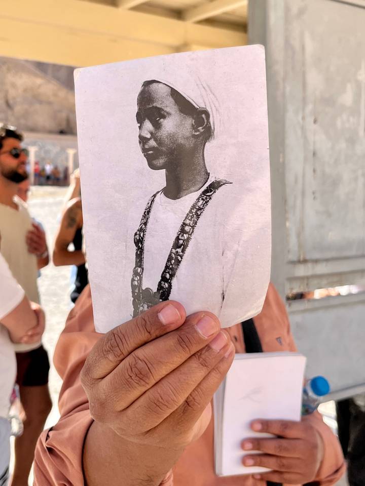 Hand holds an old black-and-white photo while blurred visitors stand in the sunny background.
