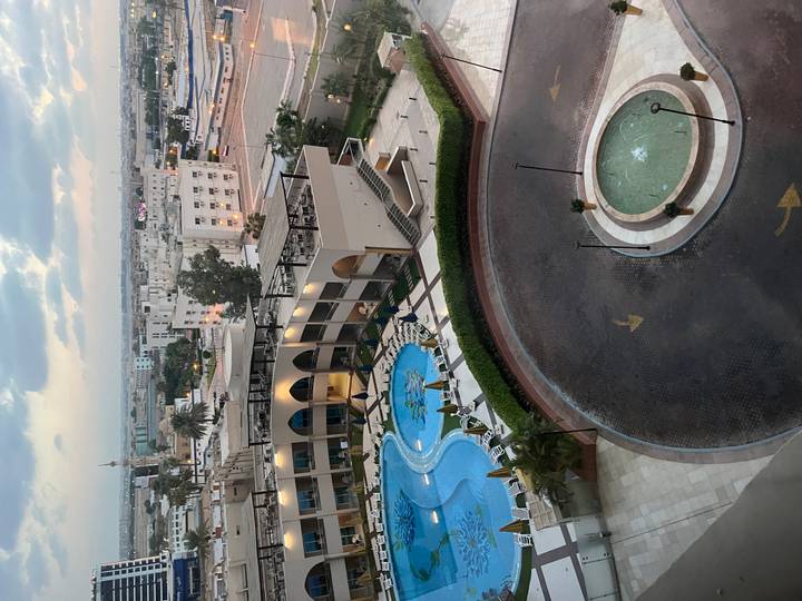 High hotel view overlooking a curving driveway, swimming pools and city buildings at dusk.
