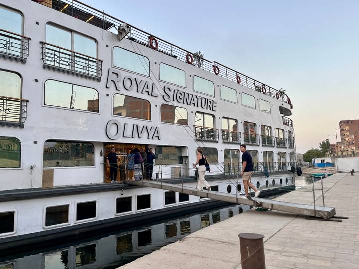 Passengers walk a gangway onto the white Nile cruise boat 'Royal Signature OliyyA' at sunset.