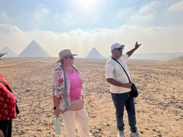 Guide gestures toward distant pyramids while explaining to a traveller in the Giza desert.