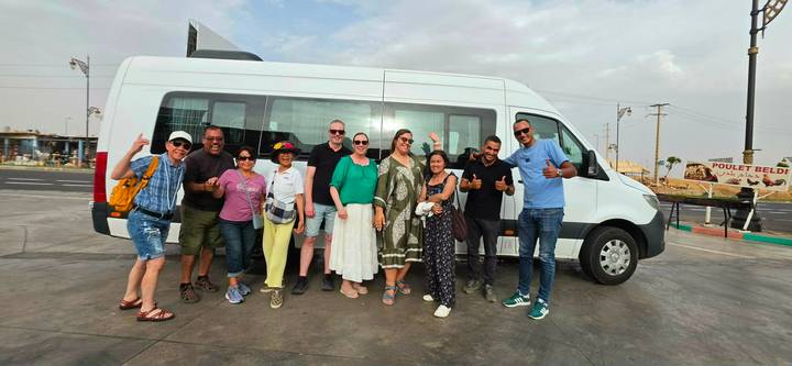 Tour group standing in front of their white minibus giving thumbs up in a parking lot.