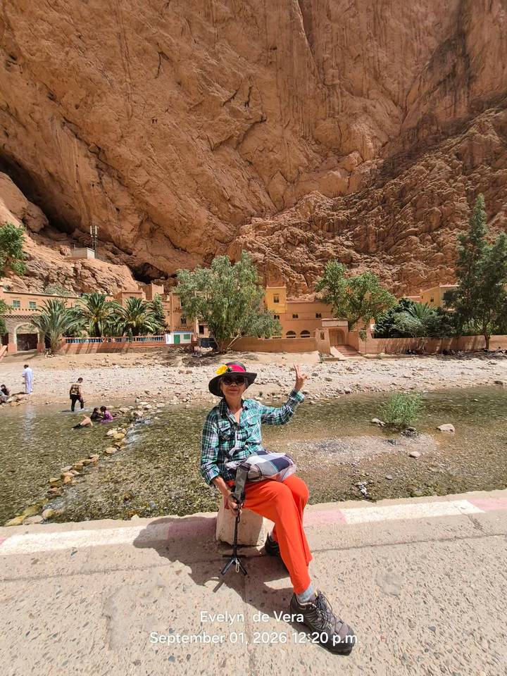 Woman waving from a shallow stream with towering orange cliffs of Todra Gorge behind.