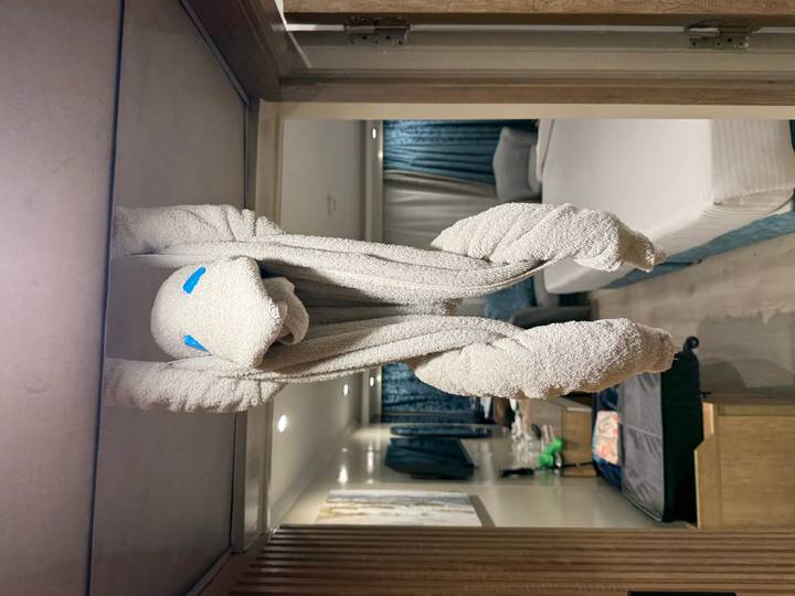 A creatively folded towel animal hanging from a shelf in a neat cruise-style cabin with a bed and furnishings in the background.