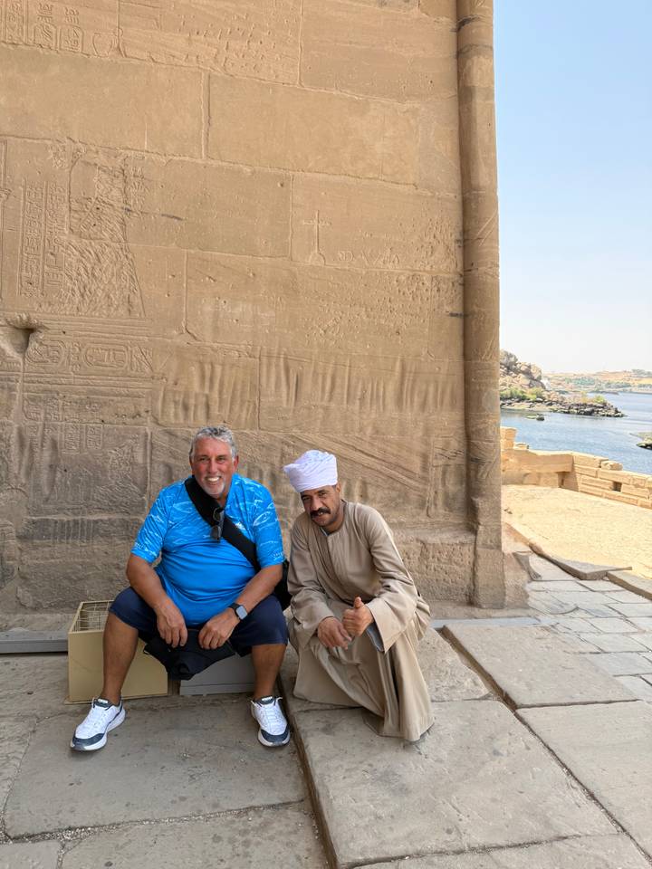 Two men sit and smile beside an ancient sandstone wall covered in hieroglyphs with the Nile visible in the distance.