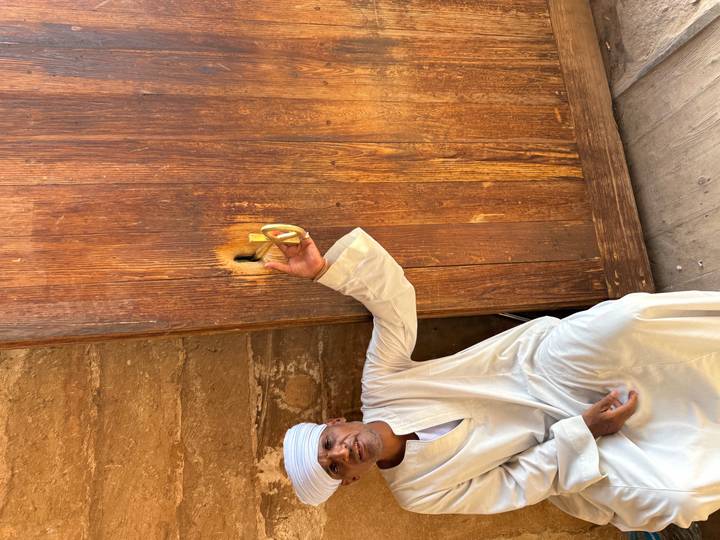 An Egyptian man in white galabeya points at a traditional wooden lock on a large rustic door.