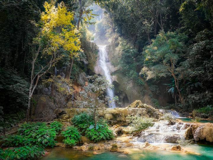Multi-tiered Kuang Si waterfall cascades through dense jungle into turquoise pools surrounded by greenery.