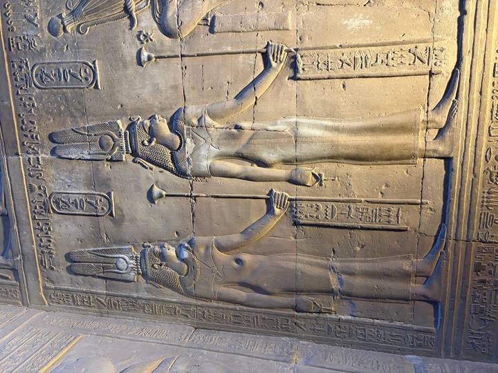 Stone reliefs of Egyptian goddesses holding scepters, illuminated inside a temple chamber.
