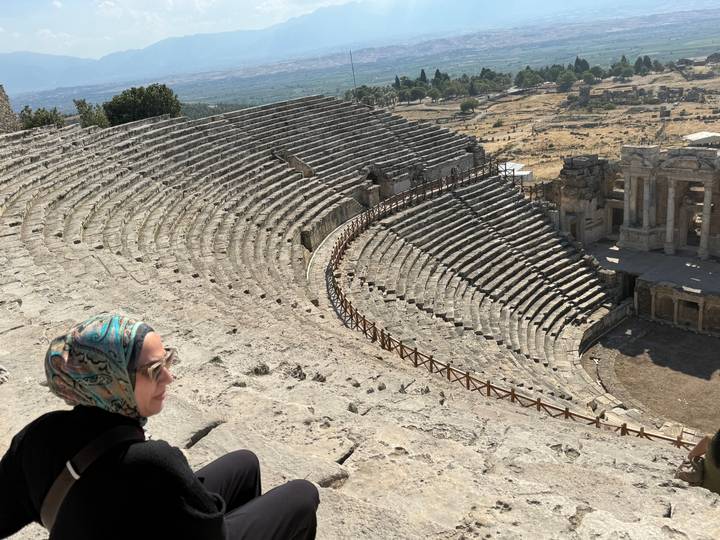 A visitor in a headscarf sits overlooking the sweeping stone tiers of an ancient Roman amphitheatre set on a dry hillside.
