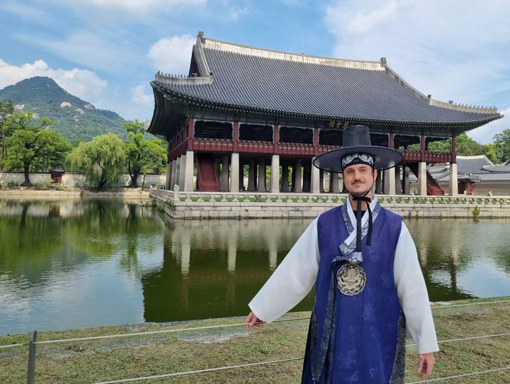 Man in traditional hanbok standing before Gyeongbokgung Palace pavilion reflected in a pond
