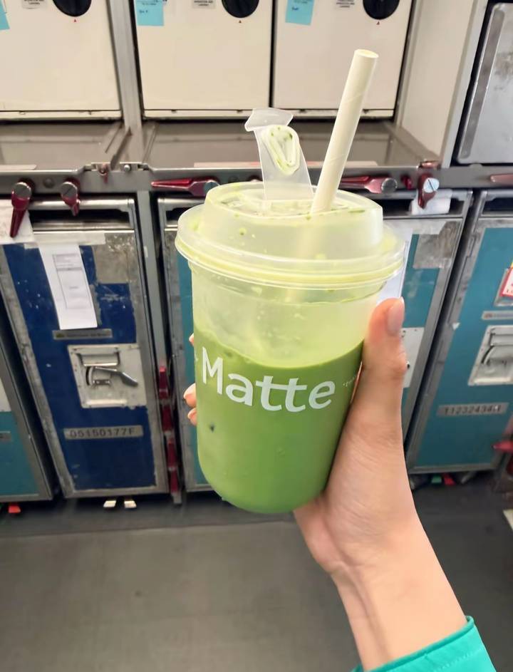 Hand holding a plastic cup of green iced matcha drink in front of metal airline service trolleys