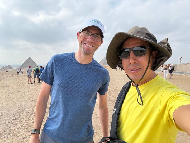 Two travelers take a selfie with the pyramids of Giza visible behind them on a hazy day.