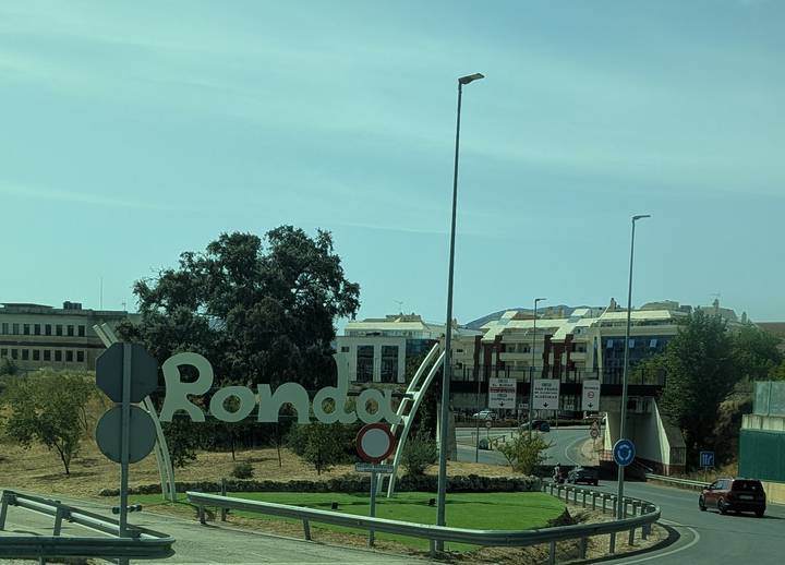 View through a bus window of a roadside sign reading 'Ronda' with modern buildings in the background.