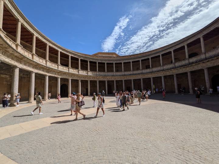 Tourists explore the circular colonnaded courtyard of the Palace of Charles V under a vivid blue sky with wispy clouds.