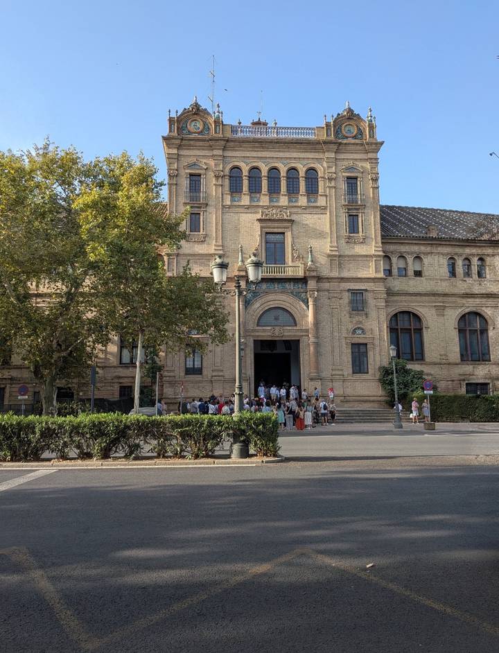 A tour group gathers at the entrance of a grand historic building framed by mature trees in Seville.