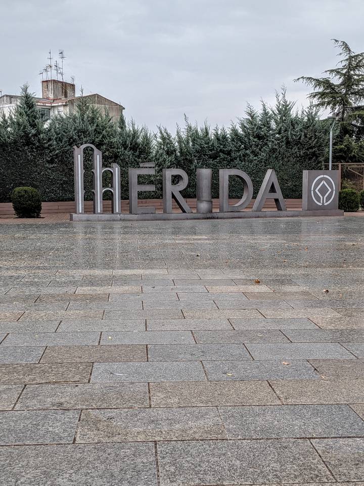 Large metal letters spelling MERIDA on a wet paved plaza with trimmed hedges behind.