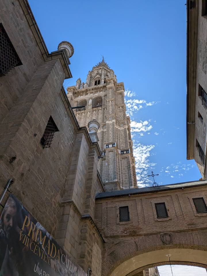 Upward view of Toledo Cathedral’s soaring bell tower framed by stone walls and a bright sky streaked with clouds.