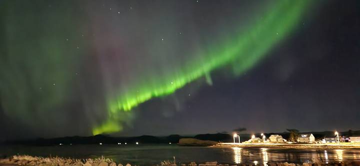 Green northern lights arc across a dark night sky above a quiet coastal village reflected in calm water.