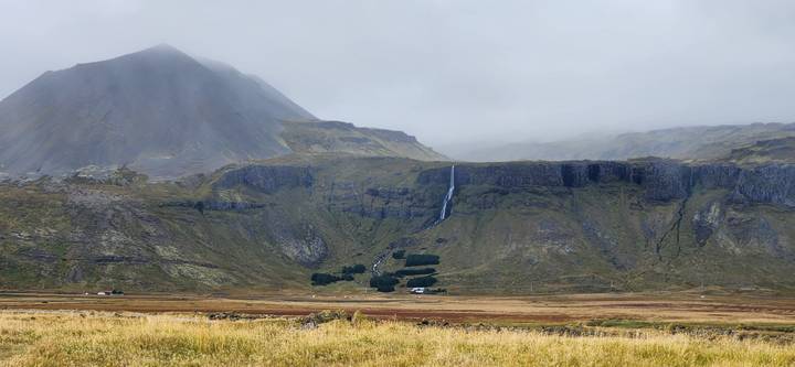 A misty mountainside with a thin waterfall dropping down steep green cliffs under an overcast sky.