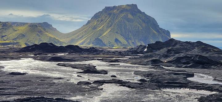 A stark black lava plain with bright streams lies before a rugged green-streaked mountain under a brooding sky.