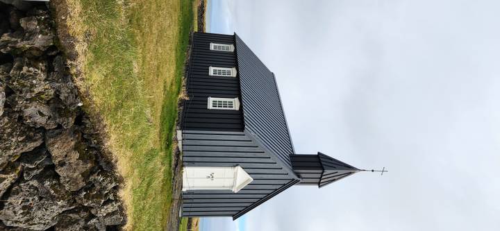 A simple black wooden church stands on a grassy lava field beneath a grey Icelandic sky.