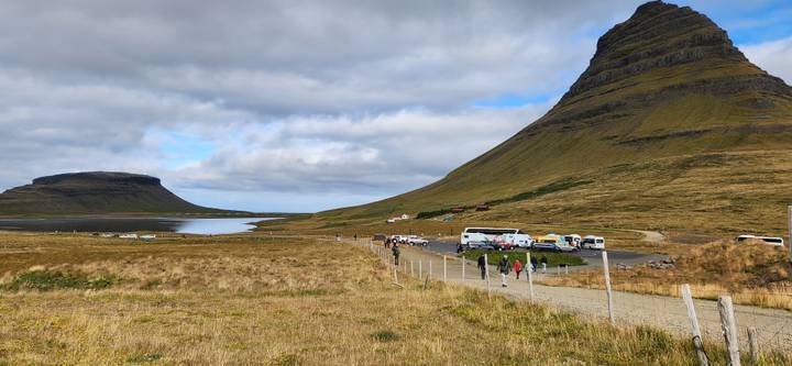 Cars and visitors gather at a viewpoint beneath the distinct cone of Kirkjufell rising over golden-brown grasslands.