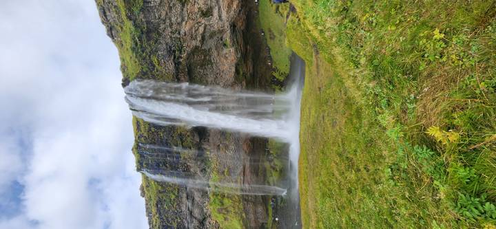 The tall veil of Seljalandsfoss plunges over a mossy cliff into a green meadow under broken clouds.