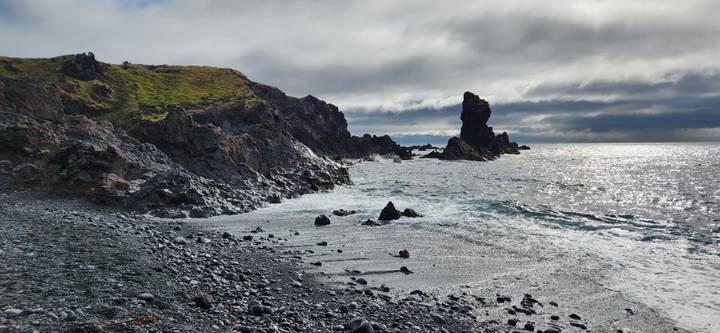 Waves crash against a rugged volcanic shoreline and a lone sea stack under dramatic clouds.