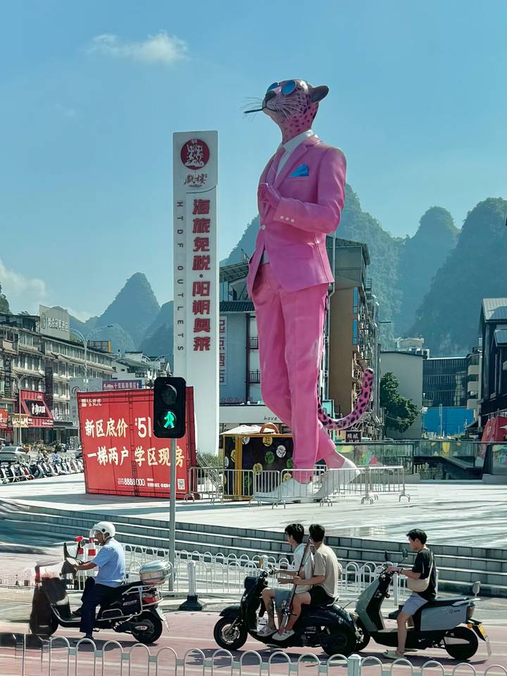 Urban scene with a towering pink-suited statue set against karst peaks, traffic lights and Chinese signage.
