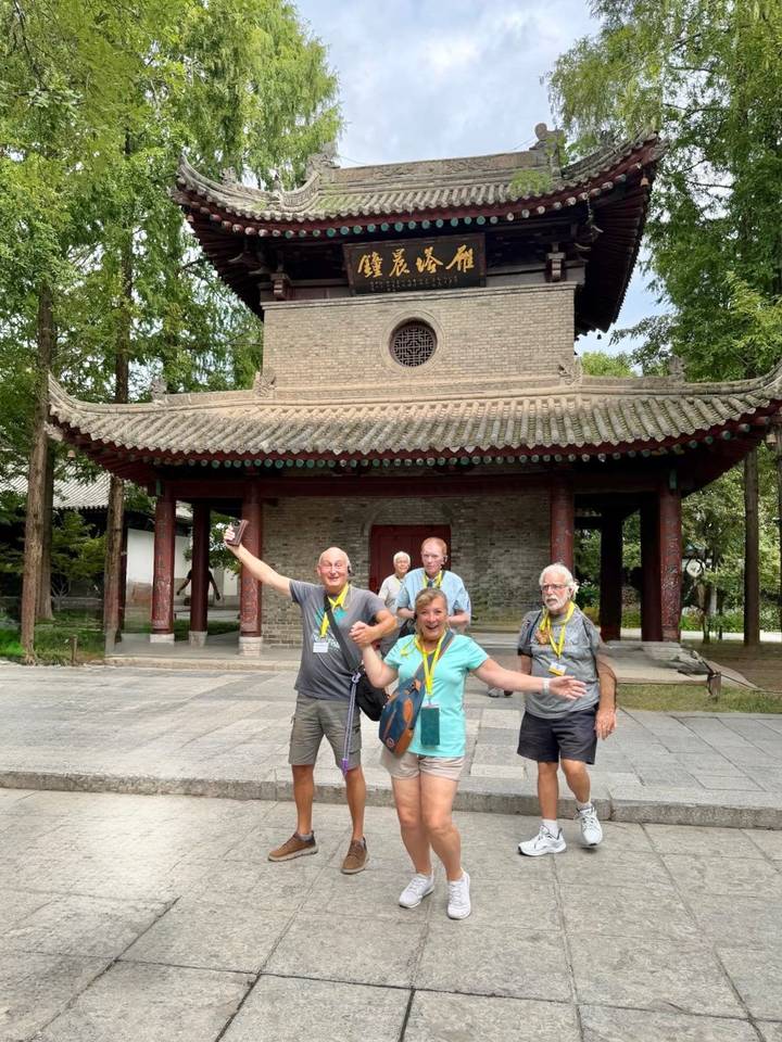 Smiling tourists pose energetically in front of an ancient tiled roof pavilion within a Chinese temple complex.