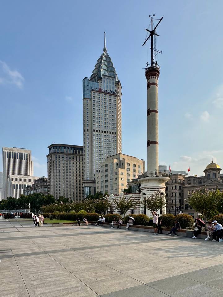 Modern high-rise buildings and a historic column stand along Shanghai’s waterfront under a clear afternoon sky.