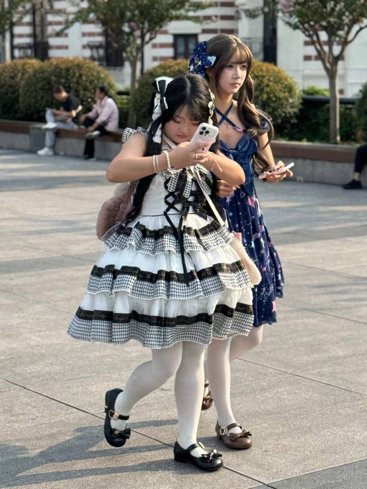 Two young women in frilly, layered dresses check their phones while standing in a sunny public square.