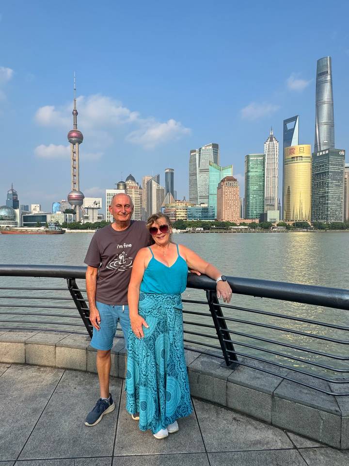 Smiling couple posing on a riverside promenade with Shanghai’s modern skyscrapers across the water under blue skies.