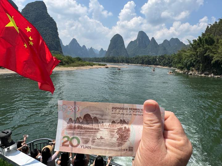 Hand holding a 20-yuan banknote that matches the karst peaks lining the Li River, with a Chinese flag fluttering on a boat.