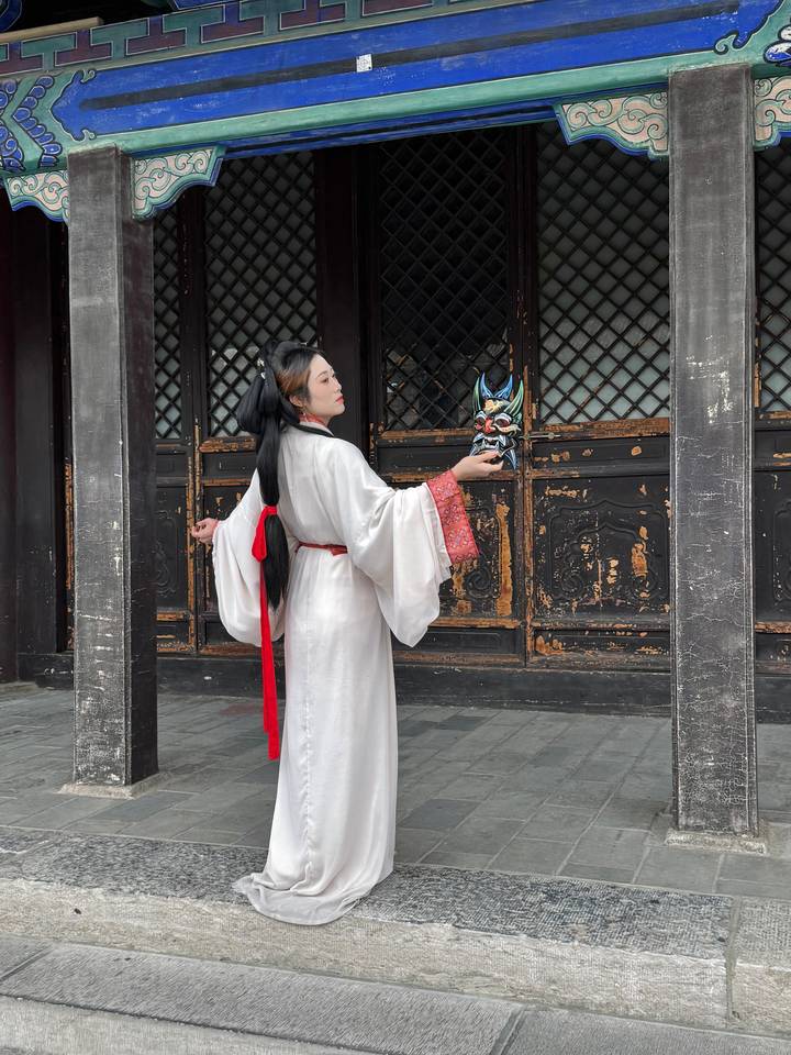 Woman in flowing white traditional costume holding a dramatic opera mask in a historic wooden courtyard.