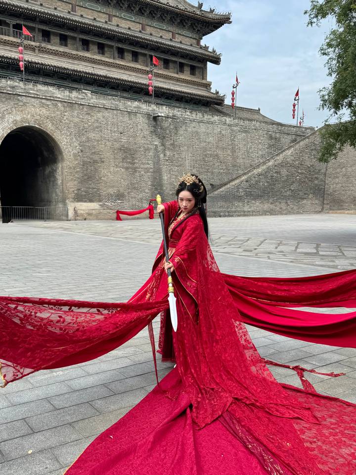 Woman in flowing red costume brandishing a sword dramatically in a large stone courtyard.