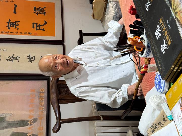 Elderly man smiling while seated at a desk filled with calligraphy tools and scrolls.