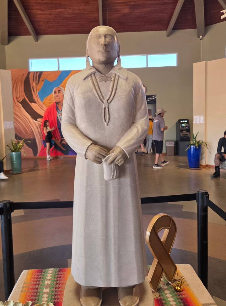 Indoor statue display of a figure in traditional attire with tourists strolling in the background.