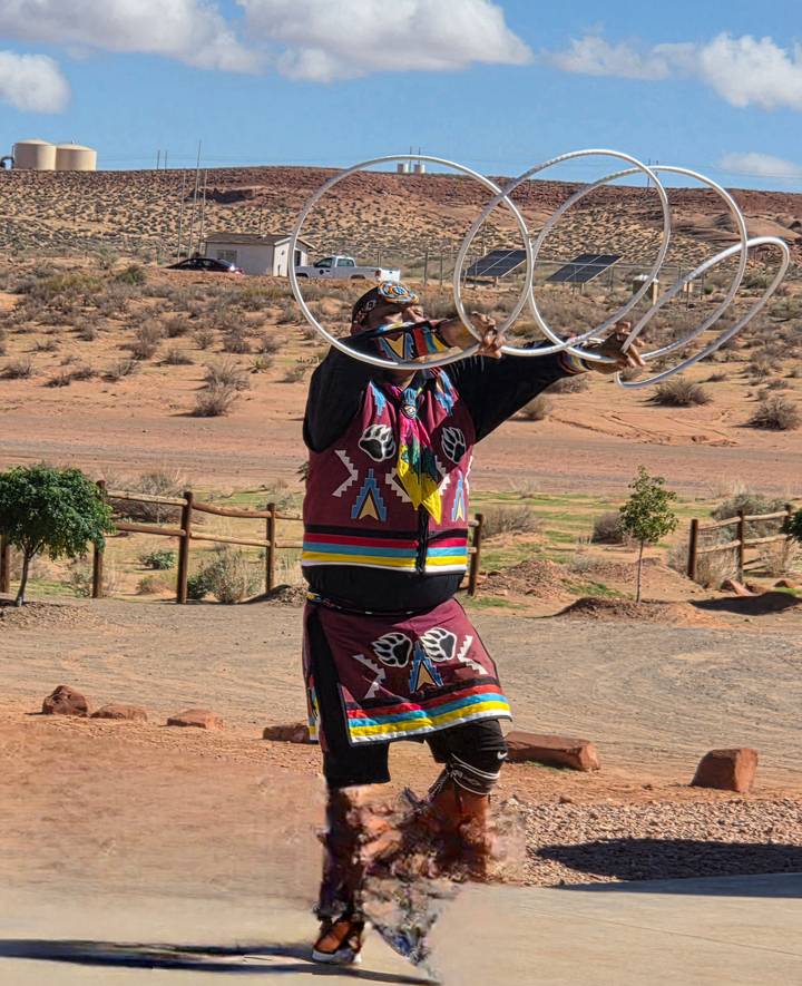 A Navajo hoop dancer performs outdoors in colorful regalia against a desert backdrop.