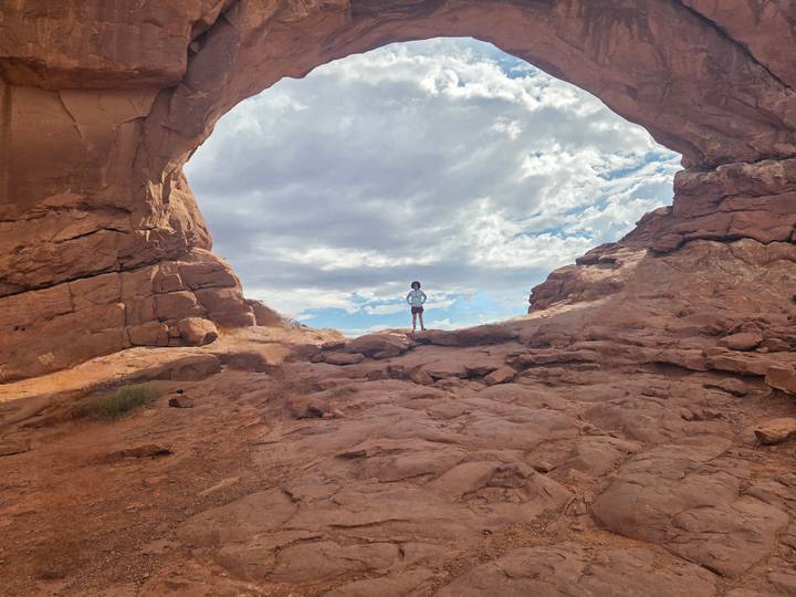 A lone hiker stands beneath a vast natural sandstone arch with dramatic clouds overhead.