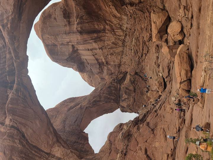 Visitors scramble over boulders beneath a pair of intersecting sandstone arches in a rugged canyon.