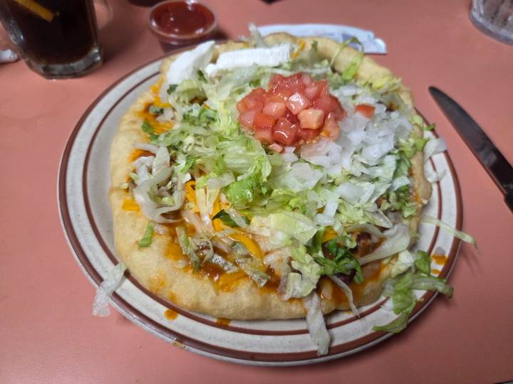 A plate of frybread topped with lettuce, cheese, onions, tomatoes and sauce sits on a diner table.
