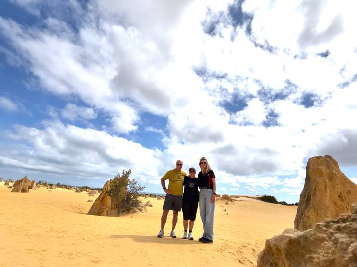 Three travellers stand among the golden limestone pinnacles of a desert landscape under a partly cloudy sky.