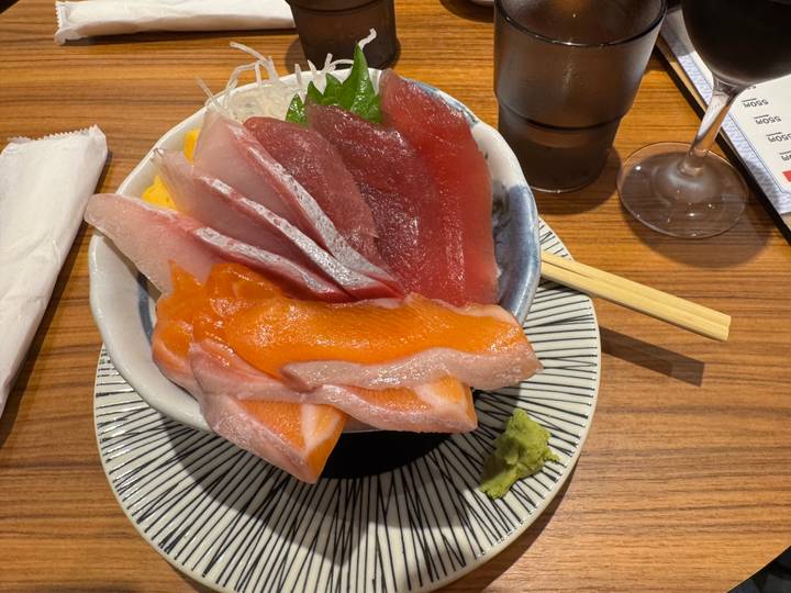 Colourful bowl of assorted sashimi slices with wasabi and chopsticks laid across a wooden counter.