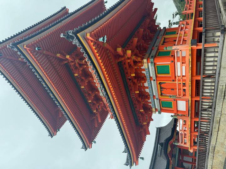 Vivid orange three-tiered pagoda with intricate wooden joinery and green panels against an overcast sky in Kyoto.