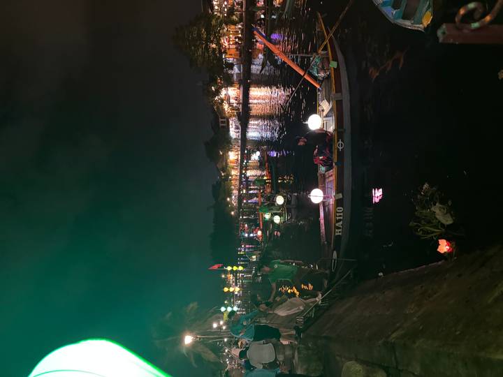 Night boats lit by lanterns glide along riverside promenade lined with glowing shops
