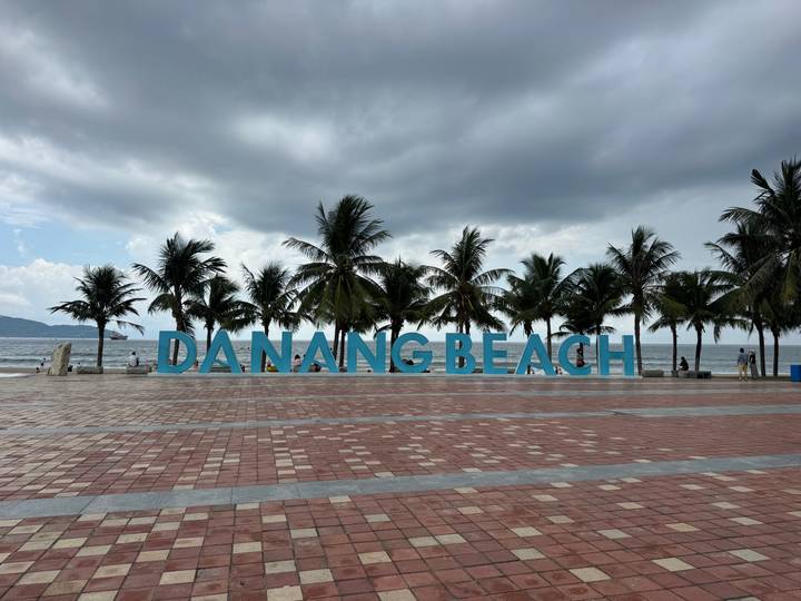 Large turquoise DANANG BEACH sign with palm trees and cloudy skies on seaside promenade